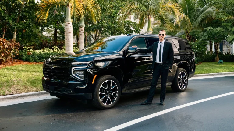 ART chauffeur in a black suit standing beside a Chevrolet Suburban in a palm-lined driveway