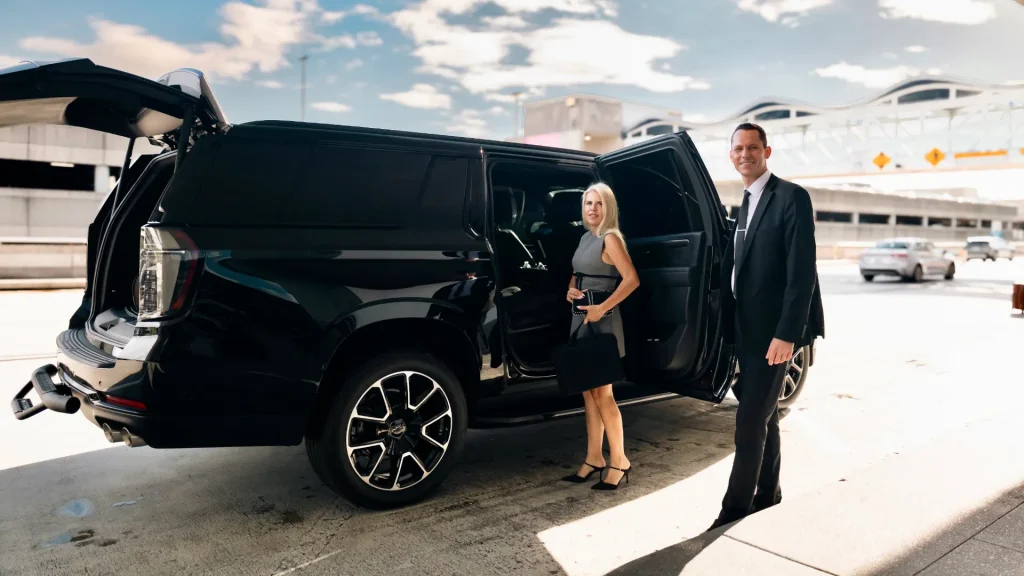 ART chauffeur in a suit beside a black Suburban with a passenger and her luggage