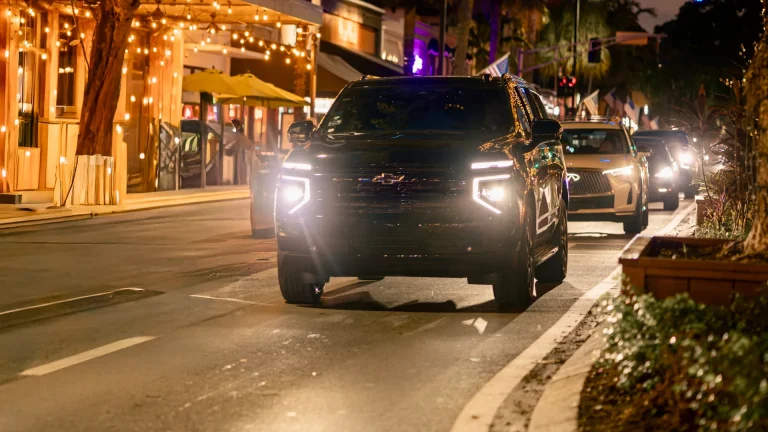 Black Suburban with headlights on, driving down a string-lit street at night
