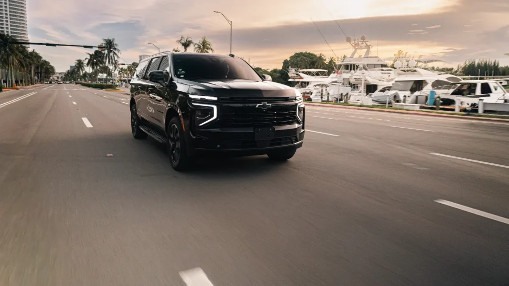 Black Suburban driving along a waterfront road at sunset