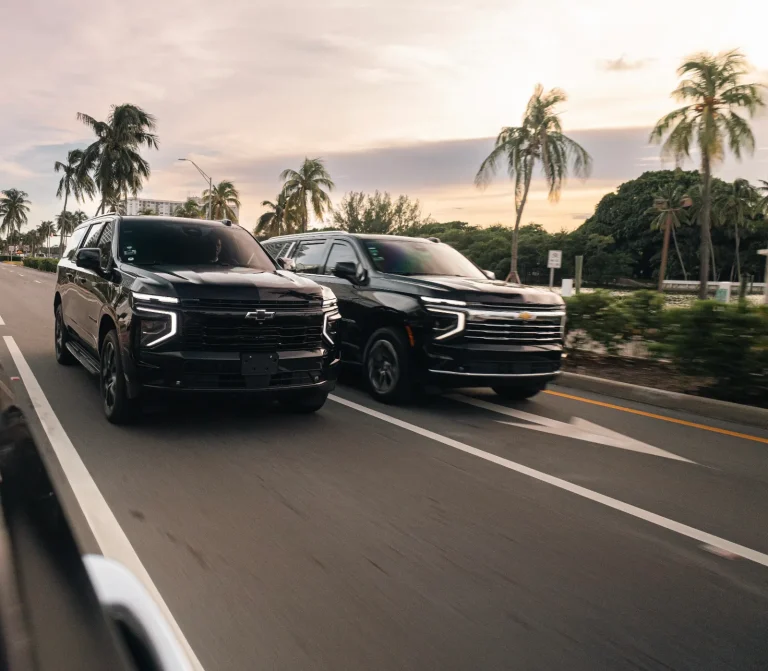Two black Chevrolet Suburbans driving side by side on a palm-lined road at sunset